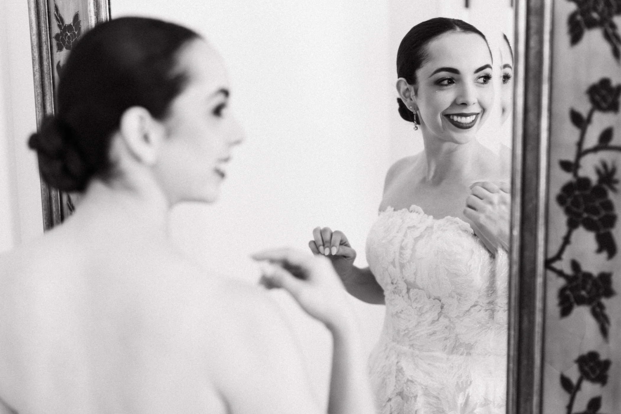Black and white photo of Paulina smiling at her reflection while putting on her earrings before the wedding