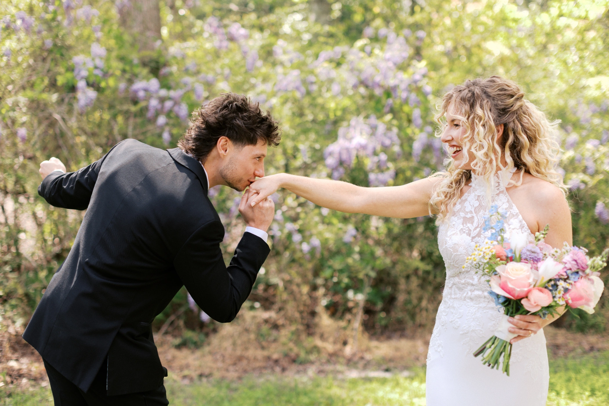 Zane kissing Taylor's hand in front of blooming wisteria during their first look portraits